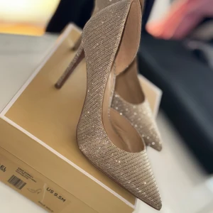 Glittriga guldiga pumps med spetsig tå - Snygga pumps i guld med glittriga detaljer och spetsig tå. Skorna har hög klack och en elegant form som verkligen sticker ut. Perfekta för dig som vill ha något extra till outfiten. Materialet är syntet med glittrig finish.