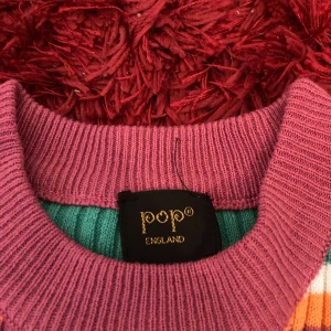 Randig ribbad tröja från Pop England - Färgglad ribbad tröja från Pop England med korta ärmar. Tröjan har gröna, lila, orangea och vita ränder samt en lila ribbad krage. Perfekt för dig som gillar retrovibbar och vill sticka ut med en unik stil.