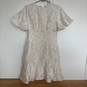 Vit studentklänning med volang - Superfin vit kortklänning från i storlek M. Klänningen har puffiga korta ärmar, blommigt mönster och volang nertill. Tillverkad i glansigt material som ger en lyxig känsla. 