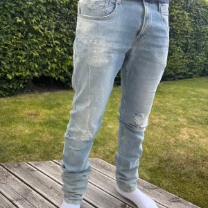 Replay Anbass ljusblå jeans herr W34 - Säljer ett par Replay Anbass jeans i ljusblå tvätt med slitningar och en trasig detalj vid knät. Klassisk femficksmodell med dragkedja och knapp. Jeansen är i bomull och har en slim passform som sitter snyggt. Perfekta för dig som gillar en avslappnad men trendig look.