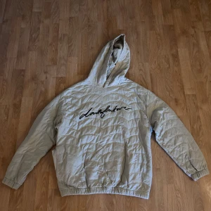 Beige quiltad hoodie från ASOS - Quiltad beige hoodue från ASOS DESIGN med huva och elastiska muddar. Stor broderad text 'darkfuture' på bröstet ger en cool vibe. Hoodien har knappdetalj vid halsen.