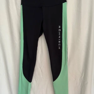 Svarta och mintgröna tights Röhnisch XS - Snygga svarta tights från Röhnisch i storlek XS med mintgröna paneler längs sidorna och nedtill på benen. Tight passform och hög midja, perfekt för träning. Stort Röhnisch-tryck på ena benet. Tillverkade i stretchigt syntetmaterial.