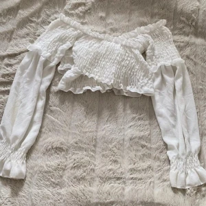 Vit offshoulder croppad blus - Superfin vit blus från Nelly med offshoulder och croppad passform. Smockad överdel och volangdetaljer längs kanterna ger en romantisk vibe. Långa ärmar med smock vid handlederna. Perfekt till jeans eller kjol för en trendig look.