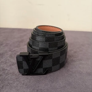 Louis Vuitton rutig svart bälte - Snyggt bälte från Louis Vuitton med klassiskt rutigt mönster i svart och grått. Bältet har en mattsvart LV-logga som spänne och är gjort i skinn med beige insida. Perfekt accessoar för att lyfta din outfit med en lyxig touch.