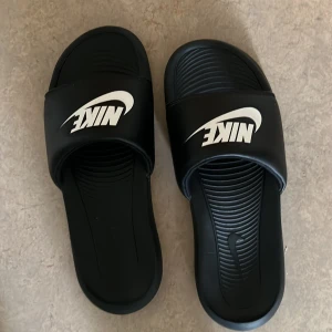 Svarta Nike sandaler med vit logga - Svarta sandaler från Nike med klassisk vit logga på ovandelen. Skorna har en bred rem över foten och räfflad sula för extra grepp. Tillverkade i syntetmaterial och perfekta för sommaren eller efter träningen.