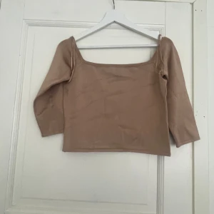 Beige topp med trekvartsärm från Guess - Snygg beige topp från Guess med trekvartsärm och fyrkantig ringning. Toppen har en liten metallbricka med Guess-logga i nacken och är gjord i mjukt material som sitter skönt på kroppen. Perfekt att styla med jeans eller kjol för en clean look. Det går sy fast metallbiten 