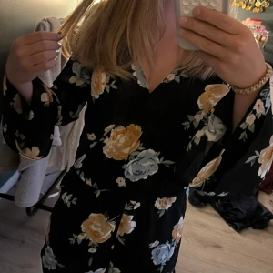 Blommig kortklänning från H&M Divided - Supersöt svart kortklänning med stora blommor i gult och blått från H&M Divided. Klänningen har v-ringning, omlott och vida trekvartsärmar med volang. Inbyggda shorts. Storlek 34