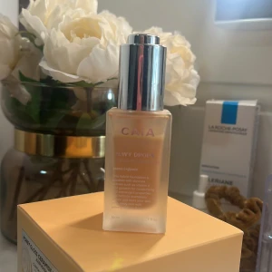 CAIA Dewy Drops Foundation 10W - Foundation från CAIA i nyansen 10W. Kommer i en snygg frostad glasflaska med silvrig pump. Ger en dewy finish och innehåller vitamin E och glycerin för extra återfuktning. Perfekt för dig som vill ha en naturlig glow och jämn hudton.