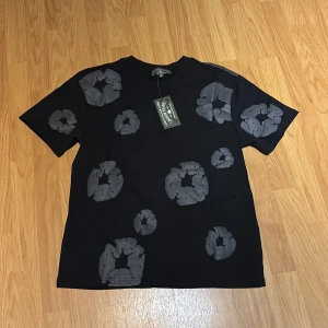 Svart Denim Tears T-Shirt  - Svart t-shirt med unika gråa blommiga mönster över hela plagget. Klassisk passform med rund hals och korta ärmar. Materialet känns mjukt och skönt, perfekt för dig som vill sticka ut med en cool och modern stil.