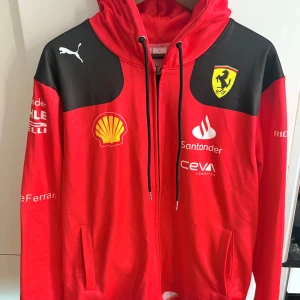 Röd Ferrari Puma hoodiejacka M - Snygg röd hoodiejacka från Puma och Ferrari med svarta axelpartier, dragkedja och huva. Jackan har flera tryckta loggor som Shell, Santander och Ferrari-emblem på bröstet. Perfekt för dig som älskar motorsport och vill sticka ut.