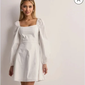 Student klänning - Superfin vit klänning från Nelly med fyrkantig ringning och långa puffärmar. Modellen är kort och har en lätt utställd kjol samt dragkedja bak. Perfekt för dig som gillar en clean och stilren look. Frakt tillkommer