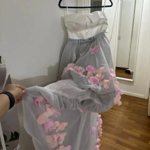 Långklänning med 3D-blommor och snörning - Drömmig långklänning med vit satintopp, snörning i ryggen och grå tyllkjol dekorerad med 3D-blommor i rosa och ljusrosa. Kjolen har volym och ett romantiskt uttryck, perfekt för dig som vill sticka ut. Smala axelband och markerad midja med rosett.