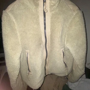 Beige teddyjacka från BDG Urban Outfitters - Säljer en beige teddyjacka från BDG Urban Outfitters. Jackan har mysigt teddyfoder, dragkedja framtill och två fickor med dragkedja. Perfekt för kalla dagar och ger en chill vibe till din outfit.