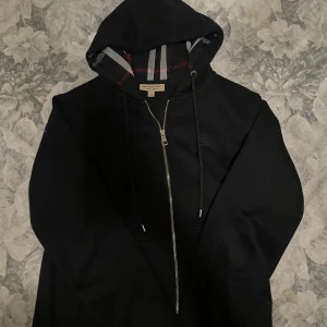 Svart Burberry hoodie med dragkedja - Svart hoodiejacka från Burberry med klassiskt rutigt foder i huvan. Jackan har dragkedja framtill, snörning vid huvan och diskret Burberry-logga på bröstet. Perfekt för dig som vill ha en stilren och lyxig streetwear-look. Den är i Storlek M men passar även S perfekt. ÄKTA HOODIE köpt online!!