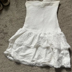 Vit kort klänning med volang - Supersöt vit kort klänning med smockad överdel och volangkjol i broderad bomull. Axelbandslös modell med fina spetsdetaljer och broderade blommor på kjolen. Perfekt för sommarens alla tillfällen!