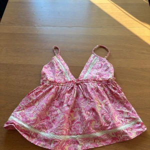 Rosa paisleymönstrad linnetopp Gina Tricot - Supersöt rosa och beige linnetopp från Gina Tricot med paisleyinspirerat mönster, smala axelband och v-ringning. Toppen har knyt i midjan och dekorativa spetsdetaljer längs bysten och nederkanten. Perfekt för varma dagar och somrig vibe.