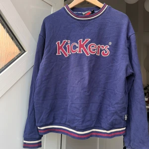 Blå Kickers sweatshirt med brodyr - Blå sweatshirt från Kickers med broderad logga i rött och vitt framtill. Tröjan har ribbade muddar och krage med röd, vit och blå rand. Långärmad och i mjuk bomull, perfekt för chill dagar. Klassisk och sportig vibe. Har ett litet hål på framsidan men inget man lägger något större märke till när den bärs! Vill få sålt den snabbt därav priset! 
