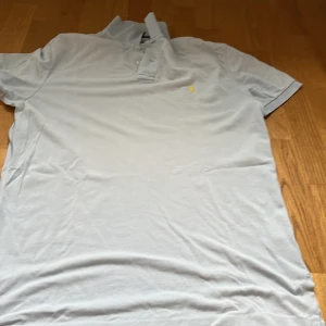 Ljusblå pikétröja från Polo Ralph Lauren - Snygg ljusblå pikétröja från Polo Ralph Lauren med klassisk krage, knäppning framtill och gul broderad logga på bröstet. Tillverkad i bomull och perfekt för en clean och stilren look. Notera att storleken är i M men den är väldigt stor - snarare L-XXL! Klicka EJ på köp nu! Har lite slitningar men det är inget man märker av!