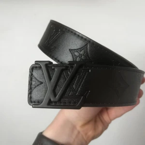 Svart Louis Vuitton skärp i skinn - Säljer ett svart Louis Vuitton bälte i skinn med präglat LV-monogram och ikoniska detaljer längs hela remmen. Bältet har en bred design och ett mattsvart LV-spänne som ger en lyxig och stilren look. Perfekt accessoar för att lyfta din outfit.
