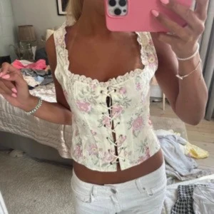 Blommig korsettopp från Gina Tricot - Supersöt korsettopp i ljusbeige med blommigt mönster i pastellfärger. Toppen har snörning framtill, spetsdetaljer längs kanterna och breda axelband. Perfekt att styla med jeans eller kjol. Helt slutsåld! (Intressekoll) aldrig använd 