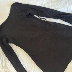 Svart långärmad klänning - Svart långärmad klänning med omlottkänsla och knytband i ryggen. Klänningen har en lätt strukturerad, krinklad yta och en djupare rundad ringning. Sitter snyggt och är perfekt för dig som gillar en stilren look. Aldrig använd