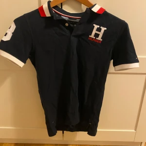 Mörkblå piké från Tommy Hilfiger - Snygg mörkblå pikétröja från Tommy Hilfiger med vit och röd detalj på kragen, broderad H-logga på bröstet och vit 8:a på ärmen. Klassisk kortärmad modell i bomull med vita muddar vid ärmslut. Perfekt för en stilren och sportig look.