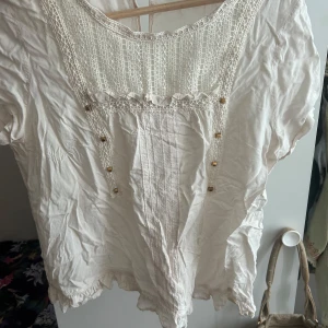 Vit blus med spets och guldknappar - Söt vit blus med spetsdetaljer framtill och små guldiga knappar. Blusen har korta ärmar, rund halsringning och volangkant nertill. Perfekt för dig som gillar romantisk och bohemisk stil. Stängning med knapp i nacken.