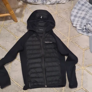 Svart pufferjacka Peak Performance S - Svart pufferjacka från Peak Performance med huva och hel dragkedja. Jackan har quiltad framsida och rygg, släta ärmar och diskret logga på bröstet. Perfekt för kyliga dagar och snygg till streetwear-stilen.