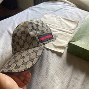 Gucci keps med GG-mönster och röd-blå rand - Exklusiv Gucci keps i beige med klassiskt GG-monogram över hela kepsen. Kepsen har en ikonisk röd och blå rand på sidan och justerbar rem bak i svart skinn. Tillverkad i Italien och levereras med originalpåse och låda.