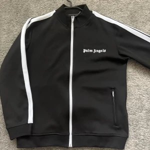 Palm Angels svart trackjacka - Svart trackjacka från Palm Angels med vita ränder längs ärmarna och vit dragkedja. Jackan har hög krage, två fickor med dragkedja och logga broderad på bröstet. Perfekt för en sportig och trendig streetstyle.