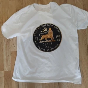 Vit t-shirt med lejontryck - Vit t-shirt med stort tryck av ett guldlejon och texten 'Empire of europe' på bröstet. Rund halsringning och klassisk passform. Perfekt för dig som vill sticka ut med ett unikt motiv.