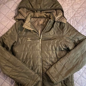 Olivgrön pufferjacka från Hummel - Snygg olivgrön pufferjacka från Hummel med huva och dragkedja framtill. Jackan har quiltad design, långa ärmar och praktiska fickor. Perfekt för kyliga dagar och enkel att matcha med olika outfits.