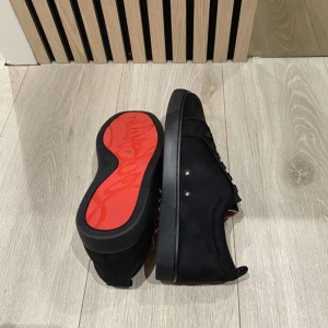  Louboutis skor - Snygga svarta sneakers från Christian Louboutin med klassisk röd sula och diskreta nitar på sidan. Skorna har rund tå, snörning och är tillverkade i mocka och skinn. Insidan är beige och de levereras med originalkartong och dustbag.