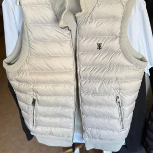 Beige dunväst från Burberry - Snygg beige dunväst från Burberry med quiltad design och ribbade kanter. Västen har två dragkedjefickor framtill, diskret Burberry-logga på bröstet och stängs med dragkedja. Perfekt för lager-på-lager och stilren look.