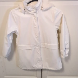 Vit regnjacka med huva och fluff inuti från Lindex - Supermjuk och fluffig vit regnkappa från Lindex med gosig fuskpäls på huvan, fram och bak. Jackan har knappar framtill, elastiska muddar vid ärmslut och en rak passform. Perfekt för regniga dagar när du vill vara både varm och snygg💞