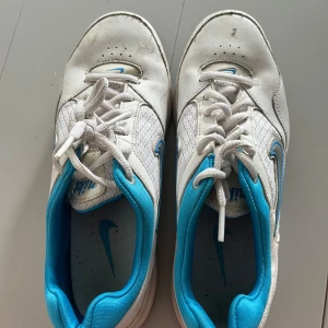 Vita och turkosa sneakers från Nike - Nike sneakers i vitt med turkosa detaljer och klassisk Swoosh-logga. Ovandel i mesh och syntet för bra andningsförmåga, med vadderad häl och platt sula. Perfekta för dig som gillar sportig stil och vill ha bekväma skor till vardagen. Storlek 37,5. 