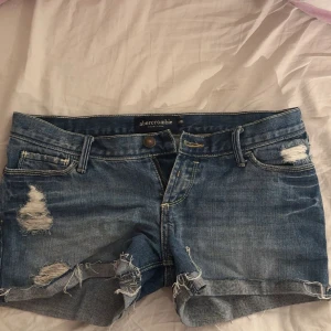 Blå lågmidjade jeansshorts från Abercrombie - Snygga blå jeansshorts från Abercrombie med slitna detaljer och fransig kant. Klassisk femficksmodell med dragkedja och knapp. Perfekta för sommardagar och ger en avslappnad vibe. De är väldigt lågmidjade och lite för små för mig så de har inte använts så ofta. Ingenting är fel på de och de har inte heller någon smutsfläck.