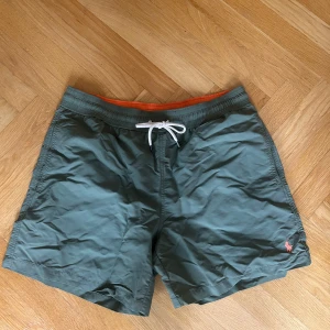 Gröna badshorts Polo Ralph Lauren M - Snygga gröna badshorts från Polo Ralph Lauren i gott skick med orange resår i midjan och vit snörning. Klassisk passform och bakficka med knapp. Perfekta för stranden eller poolen, med diskret logga broderad framtill.
