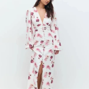 Vit långklänning med rosa blommor - Supersnygg långklänning i vitt med rosa och lila blommigt mönster. Djup v-ringning, långa vida ärmar och knappar framtill med slits. Figurnära passform i midjan och lätt glansigt material som känns lyxigt. Perfekt för sommarens alla tillfällen!