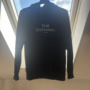 Svart hoodie från Peak Performance - Svart hoodie från Peak Performance med vit logga på bröstet. Klassisk modell med huva och långa ärmar, perfekt för en chill och sportig stil. Mjuk insida och ribbade muddar för extra komfort. 