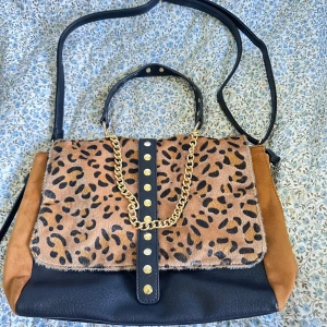Leopardmönstrad axelväska med kedja - Cool axelväska med leopardmönstrat lock, svarta och bruna partier i skinn och mocka. Guldiga nitar och kedjedetalj framtill. Väskan har både handtag och lång axelrem för olika stilar. Perfekt för dig som vill sticka ut med en trendig accessoar.