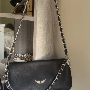Svart axelväska Zadig & Voltaire - Snygg svart axelväska från Zadig & Voltaire i skinn med silverfärgad kedjerem och ikonisk vingdetalj framtill. Väskan har dragkedja upptill, magnetlås och tryckt logga på insidan. Perfekt storlek för det viktigaste och ger en cool vibe till din outfit. Den är köpt utomlands så är nog inte äkta !💕💕💕