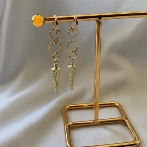 Hängande örhängen med konformad detalj - Snygga örhängen med tre ringar i rad och en konformad, nubbig detalj längst ner. Finns i både guld- och silverfärg. Ger en edgy vibe och passar perfekt om du vill sticka ut lite extra. Lätta att matcha med andra smycken.