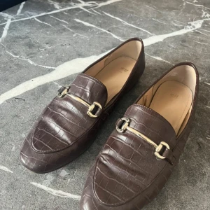 Bruna loafers med krokopräglat mönster - Snygga bruna loafers från H&M med krokopräglat mönster och guldfärgad metallspänne över vristen. Skorna har spetsig tå och är tillverkade i syntetmaterial som ger en lyxig känsla. Perfekta för att lyfta din outfit med en klassisk touch.