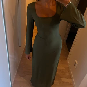 Olivgrön långärmad långklänning - Snygg olivgrön långklänning med lång ärm och fyrkantig ringning. Klänningen har en figurnära passform och är tillverkad i ett mjukt, ribbat material som sitter bekvämt på kroppen. Ryggen har korsett liknande snörning som gör att du kan anpassa hur klänningen sitter 🩷