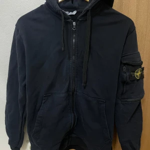 Svart hoodiejacka från Stone Island - Svart hoodiejacka från Stone Island med dragkedja, huva med snörning och klassisk logotyp på ärmen. Jackan har praktiska fickor framtill och är tillverkad i 100% bomull för en skön och avslappnad känsla. Perfekt för dig som gillar streetwear och snygga detaljer.