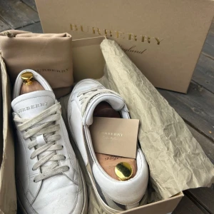 Burberry Sneakers  - Säljer ett par Burberry sneakers i vitt skinn med klassiskt rutigt tyg på sidorna. Skorna är i bra skick men diverse tecken på användning största defekten är att bokstaven ”B” har lossnat på en av skorna men detta är en liten omärkbar detalj men pga av skicket är också priset lågt. Hör gärna av er för fler bilder osv både box och dustbag följer med. Skorna passar 43
