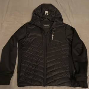 Svart pufferjacka från Sail Racing - Svart pufferjacka från Sail Racing med huva och hel dragkedja. Jackan har quiltade partier framtill och baktill, samt släta ärmar. Snygg logga på bröstet och huvan. Perfekt för kyliga dagar och riktigt stilren look.