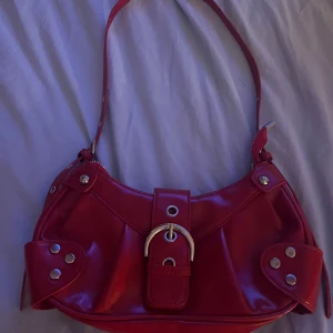 Medium stor röd axelväska - Super fin röd axelväska med silverdetaljer. Väskan är i plast men har ett läder-liknande utseende. Inte ne medium väska men lite större än en liten  – perfekt för både vardag och kväll. Lätt att matcha och bekväm att bära över axeln.  ⭐️ Stilren design med snygga silverdetaljer ⭐️ Rymlig insida med plats för det du behöver ⭐️ Lätt att bära med justerbar axelrem ⭐️ Perfekt för både vardagsbruk och utekvällar  Använd ett fåtal gånger – i mycket fint skick!