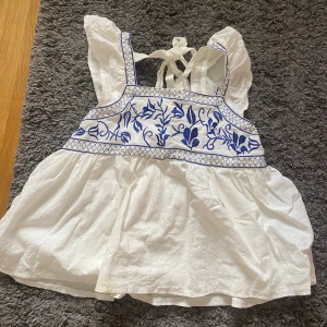 Vit blus med blå broderier och knyt - Supersöt vit blus med blå broderade blommor och rutmönster upptill. Volangiga axelband och knyt i nacke ger en romantisk vibe. Luftig och flowy modell, perfekt för varma dagar. Tillverkad i bomull för en skön känsla. Pris kan diskuteras.